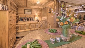 L’intérieur chaleureux d’un chalet en bois présente une table à manger en bois, dressée pour deux personnes, un lit confortable avec une literie à carreaux et un vase décoratif avec des fleurs posé sur la table.