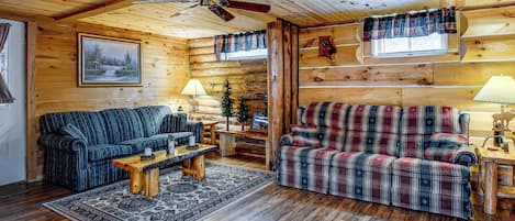 A log cabin interior with a sofa, armchair, and a small table with a lamp.