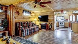A cozy cabin interior with a wooden floor, a plaid sofa, a fireplace, a television, and a kitchen area. - Arkdale Studio Cabin w/ On-site ATV Trails! (Arkdale)