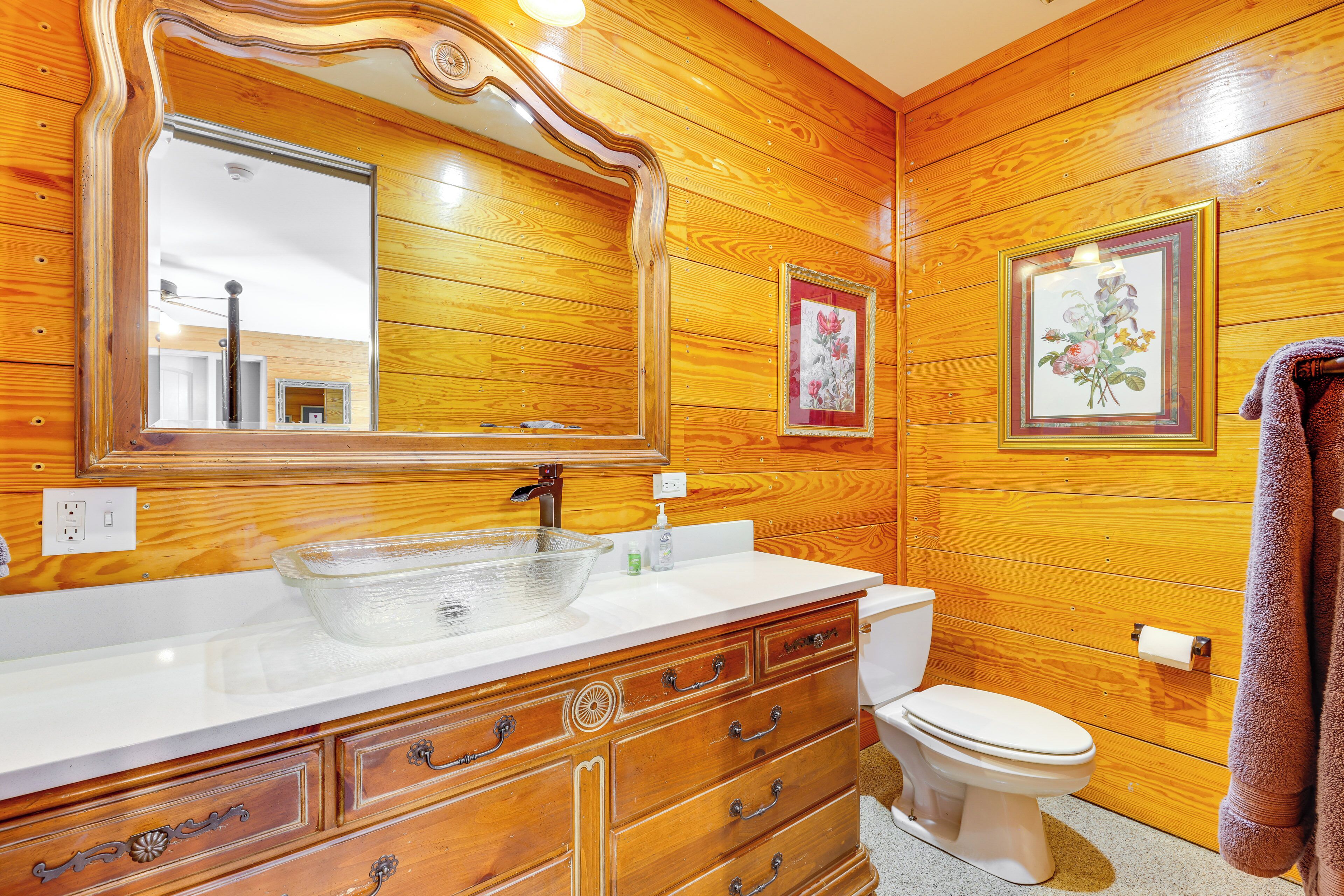 A wooden-paneled bathroom with a large mirror, a glass bowl sink, a white toilet, and a framed floral picture on the wall.