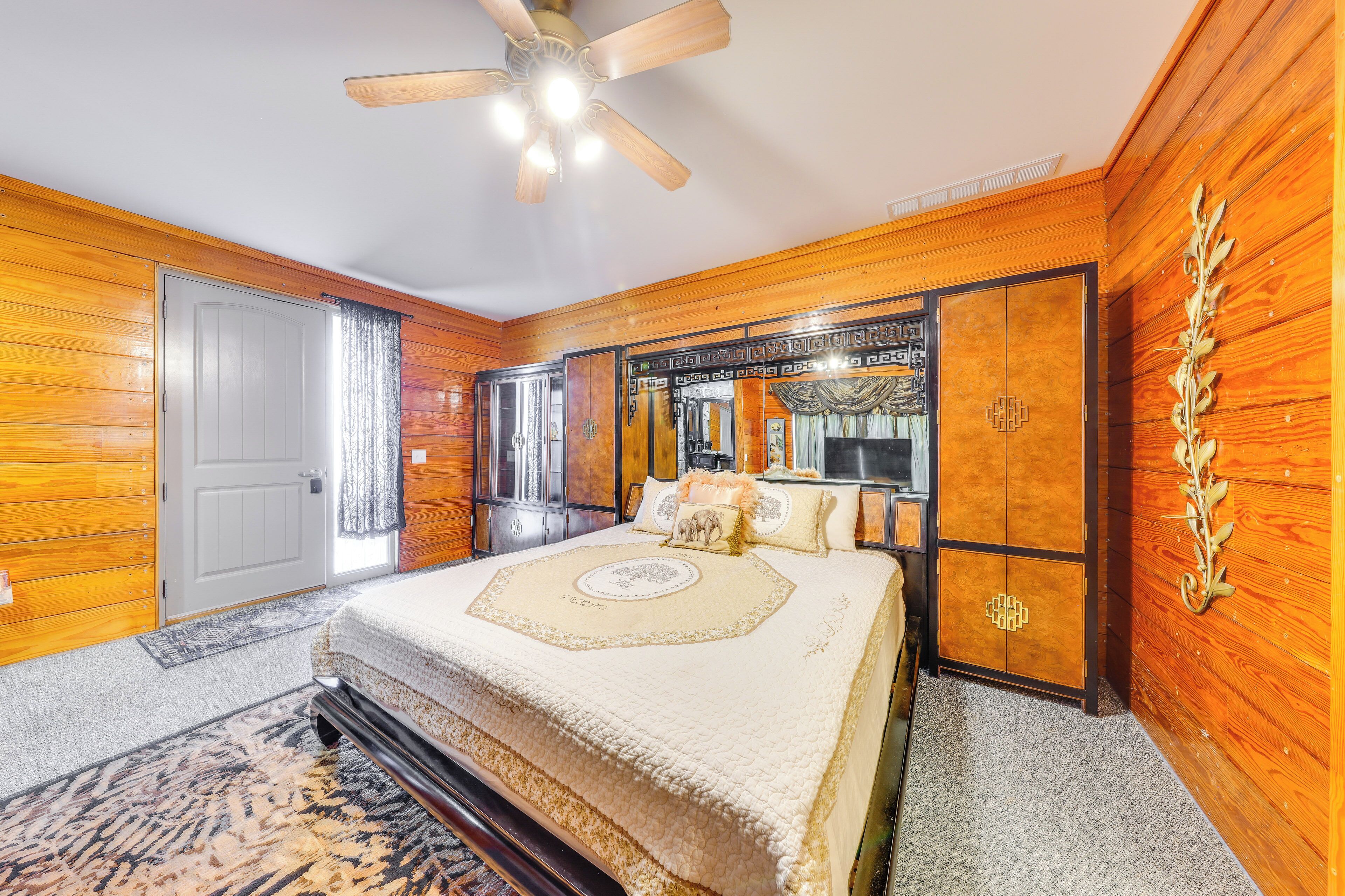 Un dormitorio con paneles de madera, una cama grande con cabecera decorativa, un ventilador de techo y una ventana con cortinas.