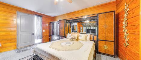 Un dormitorio con paneles de madera, una cama grande con cabecera decorativa, un ventilador de techo y una ventana con cortinas.