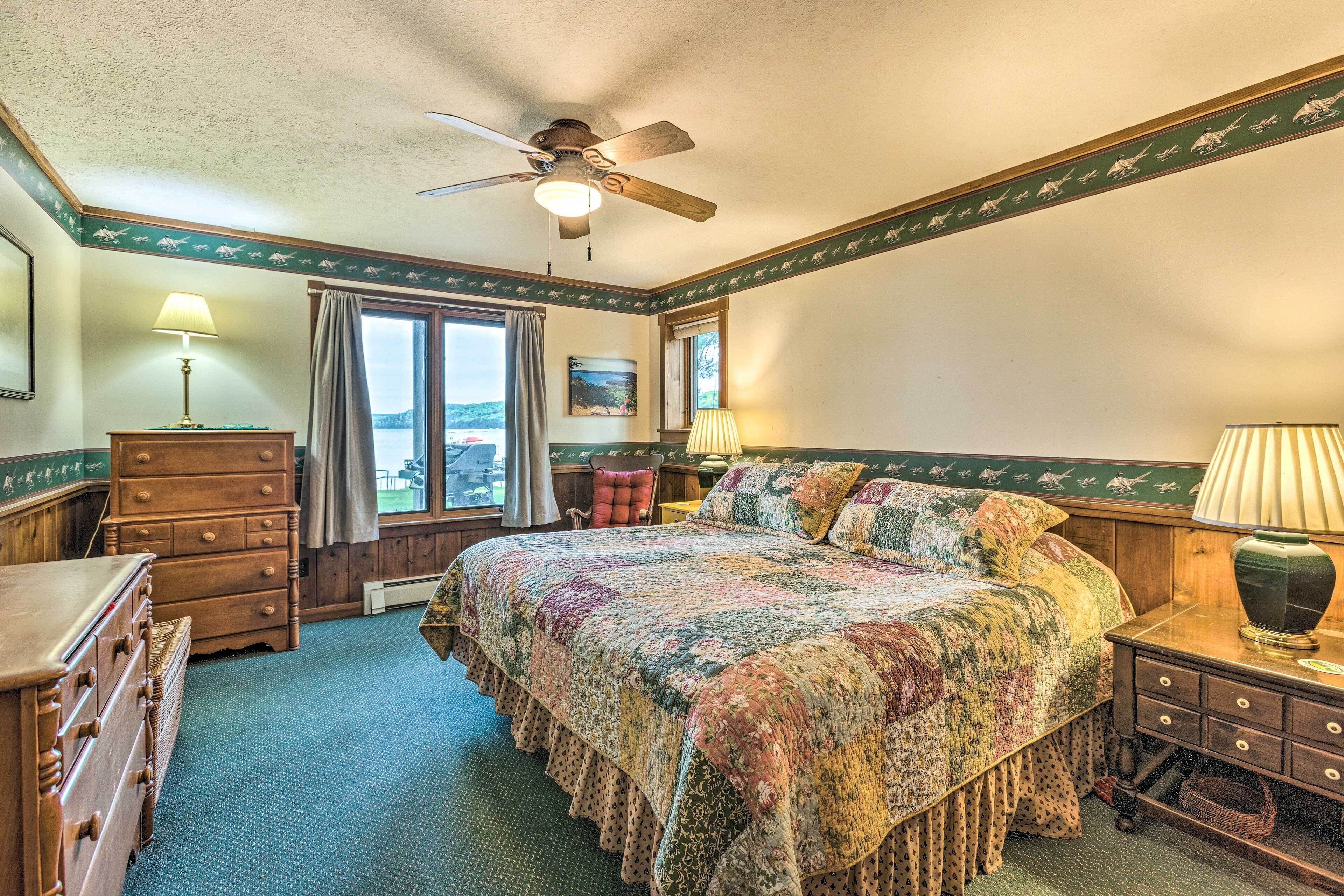 A bedroom with a large bed, a ceiling fan, a dresser, a nightstand, and a window with a view.