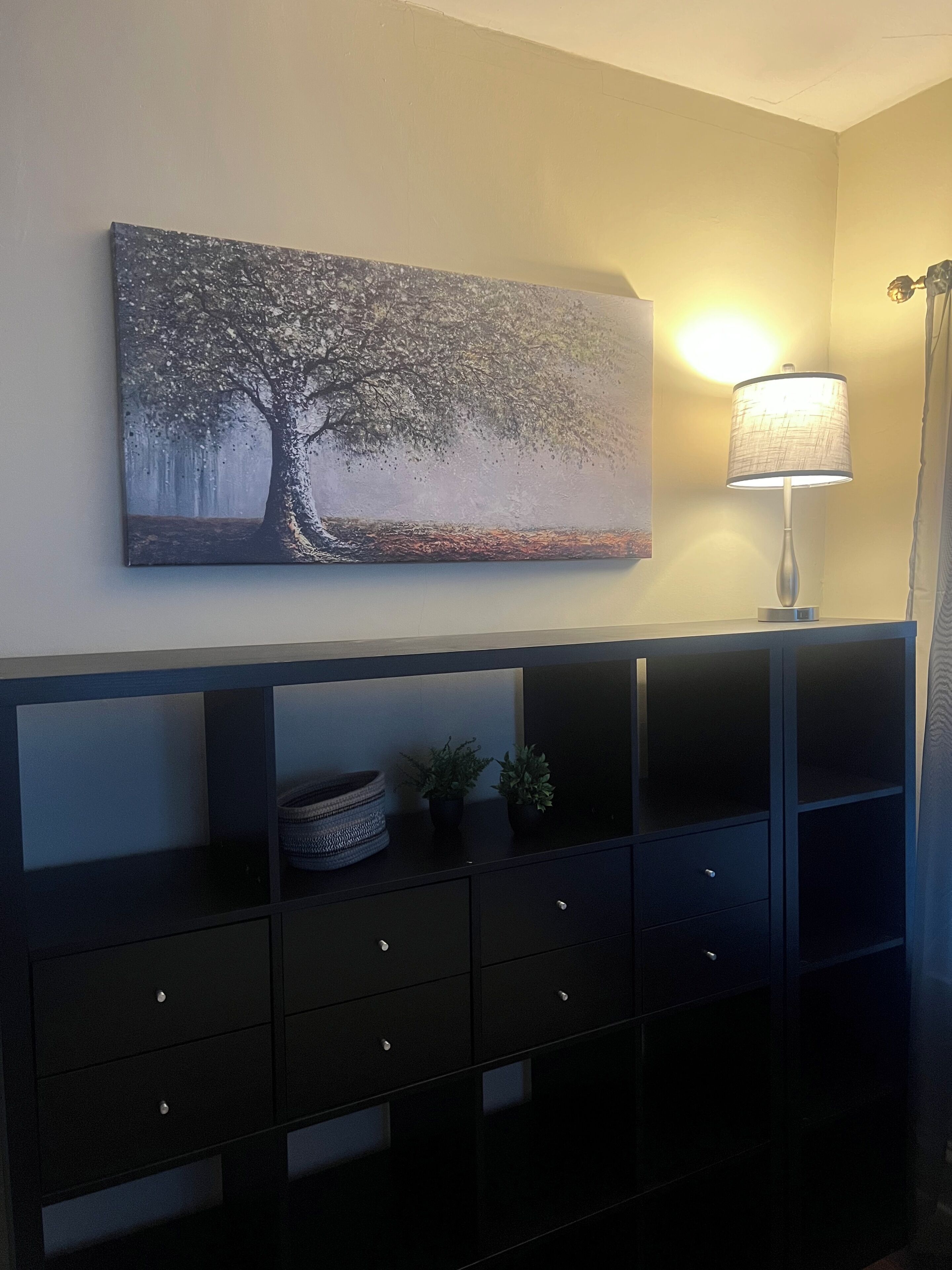 Quarto com pintura de uma árvore na parede, luminária de chão e console preto com gavetas e cesto.