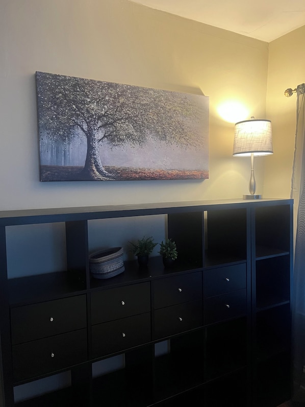 A room with a wall-mounted painting of a tree, a floor lamp, and a black console with drawers and a basket.