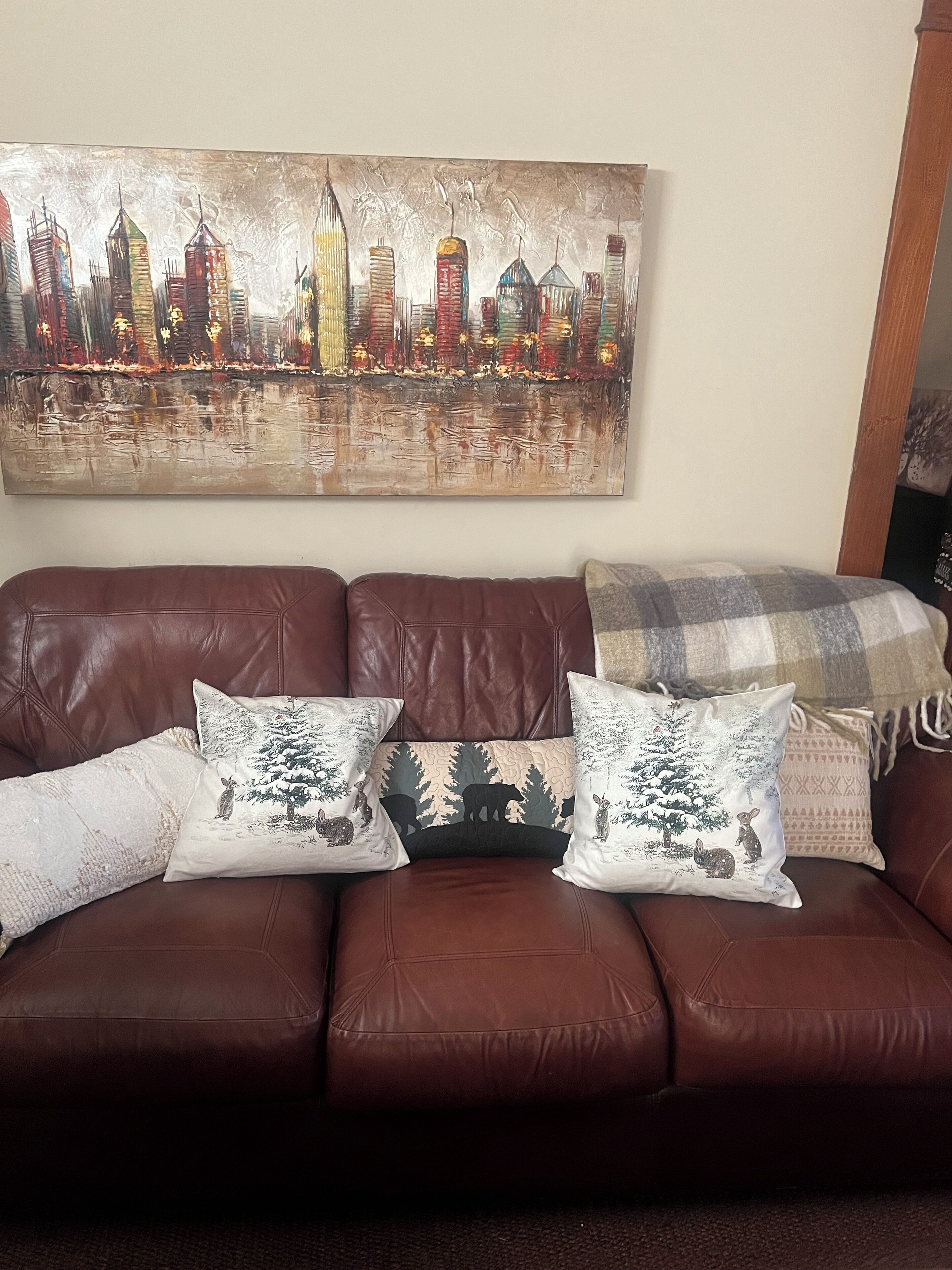 A brown leather sofa with three decorative pillows, a cityscape painting on the wall, and a patterned throw blanket.