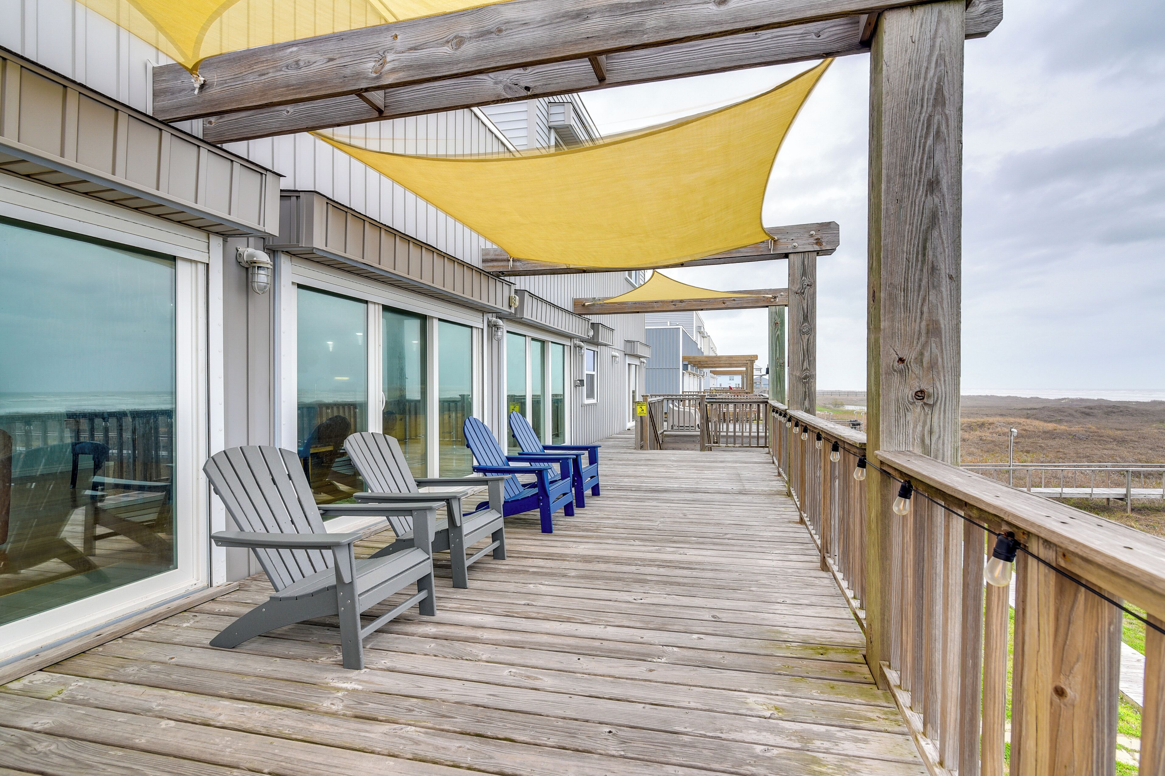 A wooden deck with Adirondack chairs, a wooden railing, and a view of the beach.