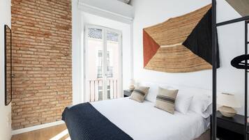 A bedroom with a brick wall, a bed with white bedding, and a geometric wall hanging.