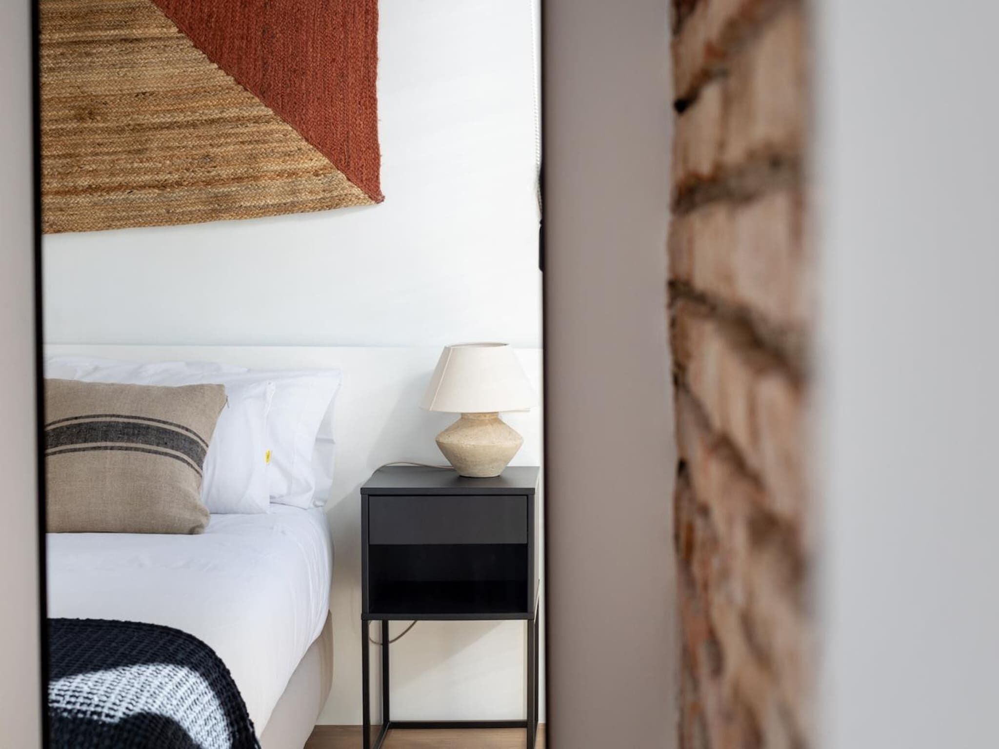 Una camera da letto con un letto, cuscini, un comodino con una lampada e un tappeto fissato al muro.