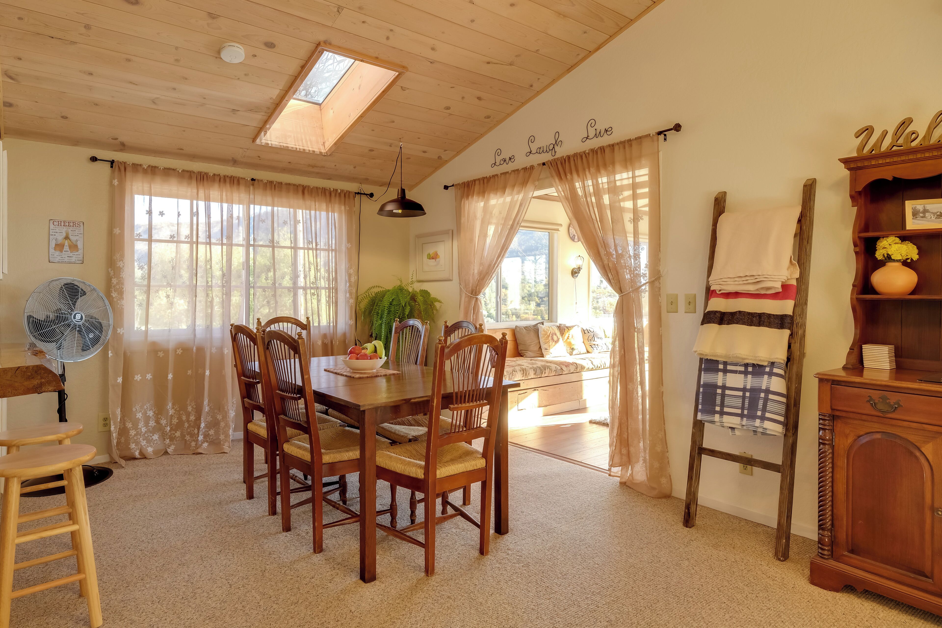 A cozy dining area with a wooden table and chairs, a fan, and a view of the outdoors through large windows.