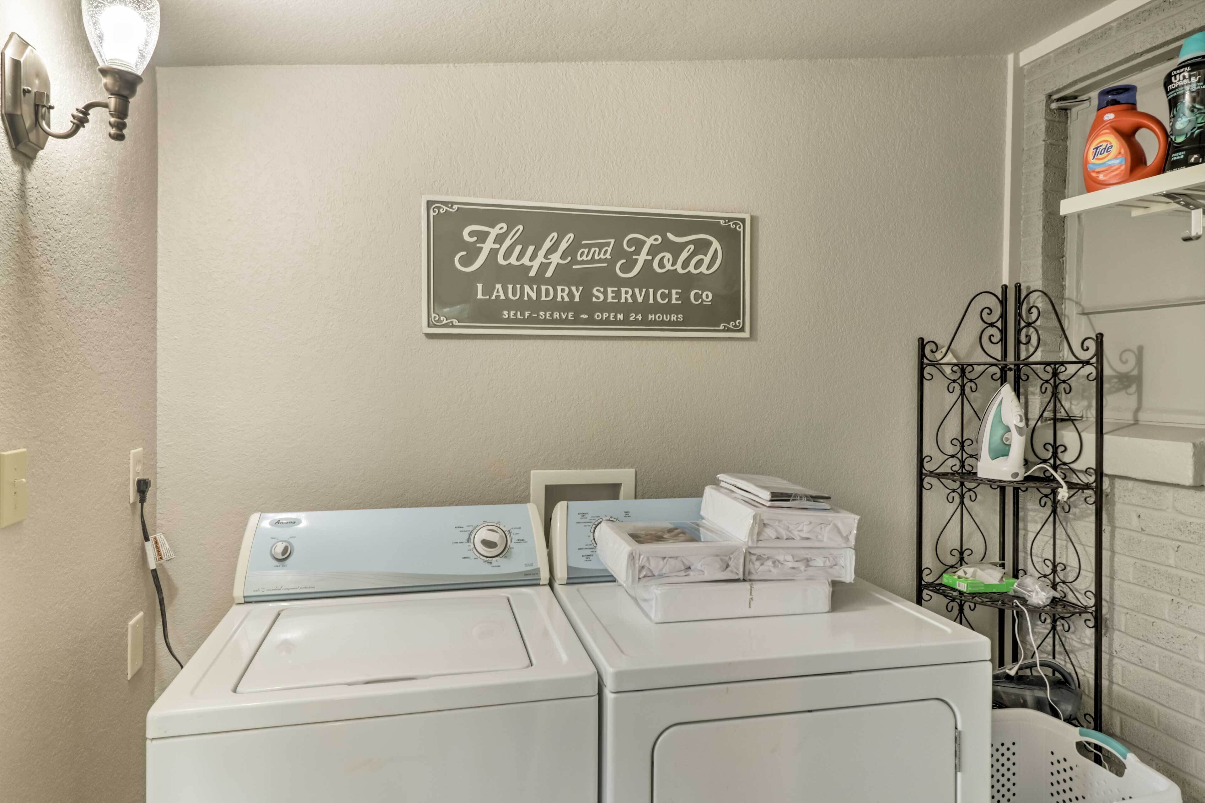 A laundry room with two front-loading washing machines, a wall-mounted sign reading "Fluff and Fold Laundry Service Co", and a metal rack with laundry supplies.