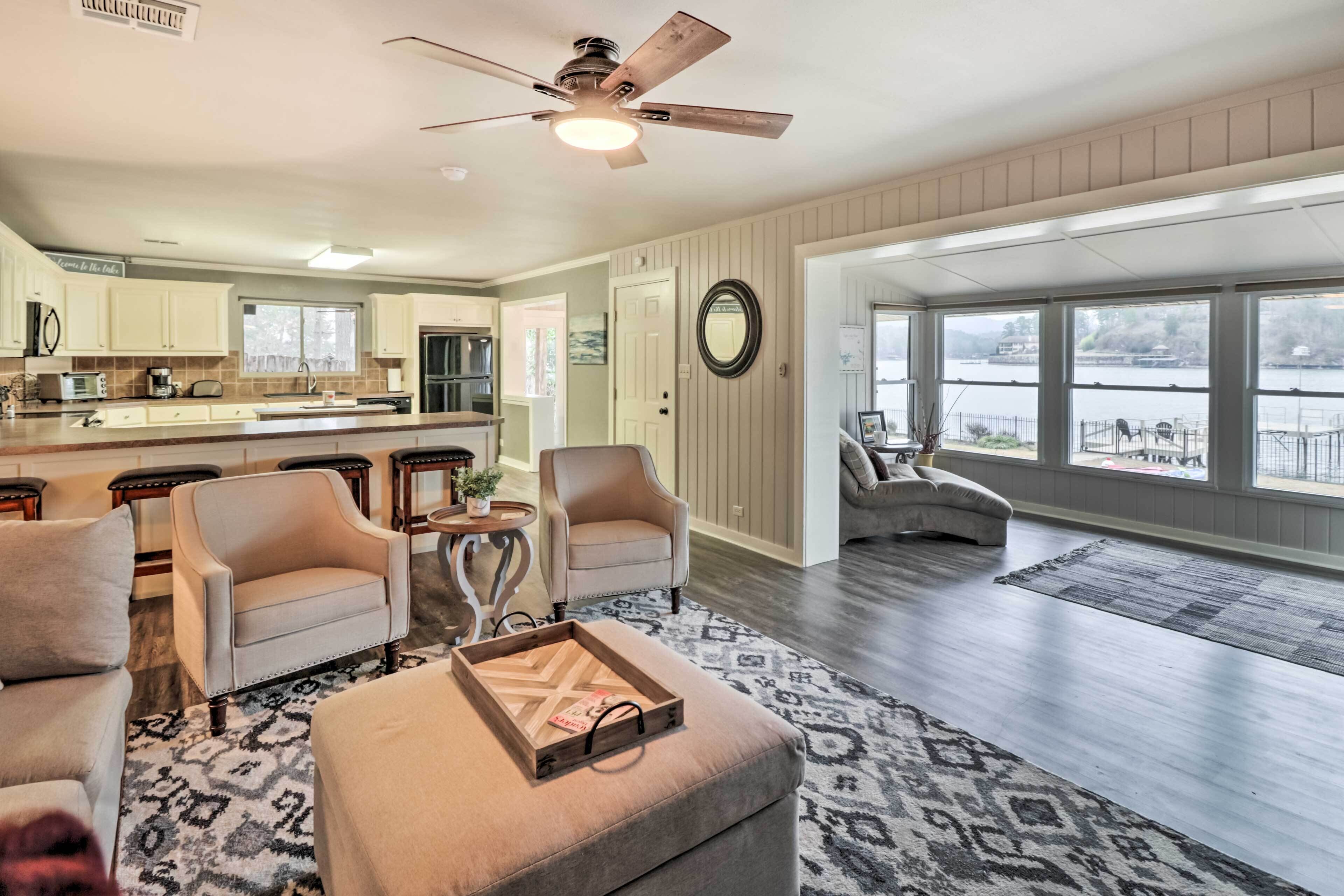 A spacious living room with a large window, a ceiling fan, and a kitchen in the background.