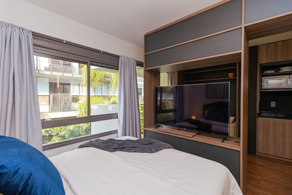 A modern hotel room with a large bed, a TV mounted on a wall, and a view of the outside balcony.