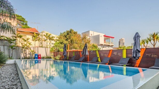 A swimming pool with lounge chairs and umbrellas, surrounded by a low wall and palm trees. - Vossa Bossa Vila Madalena Diseño (São Paulo)