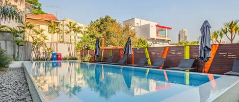 A swimming pool with lounge chairs and umbrellas, surrounded by a low wall and palm trees.
