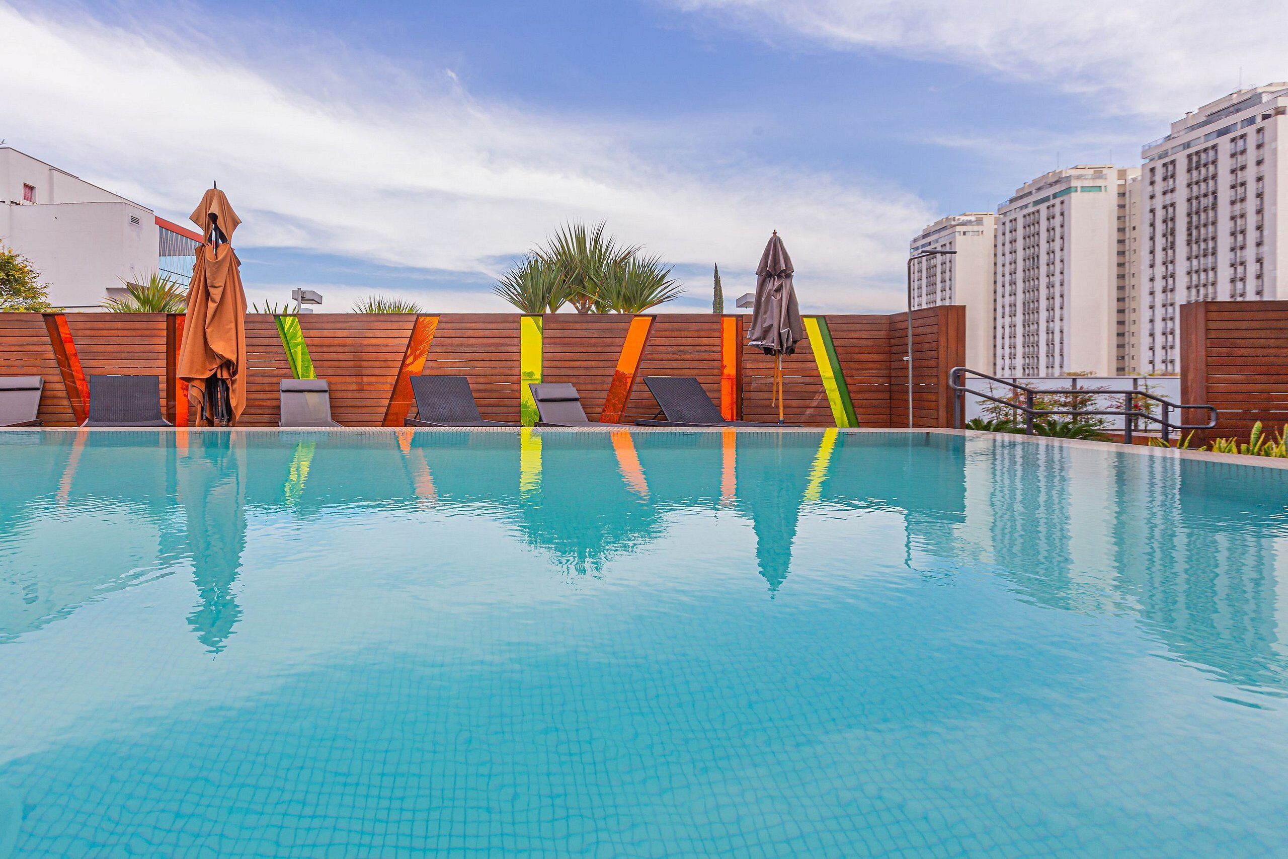 Piscina com espreguiçadeiras e área coberta, cercada por um muro de madeira e edifícios altos ao fundo.