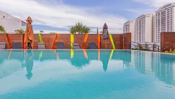 Piscina com espreguiçadeiras e área coberta, cercada por um muro de madeira e edifícios altos ao fundo.