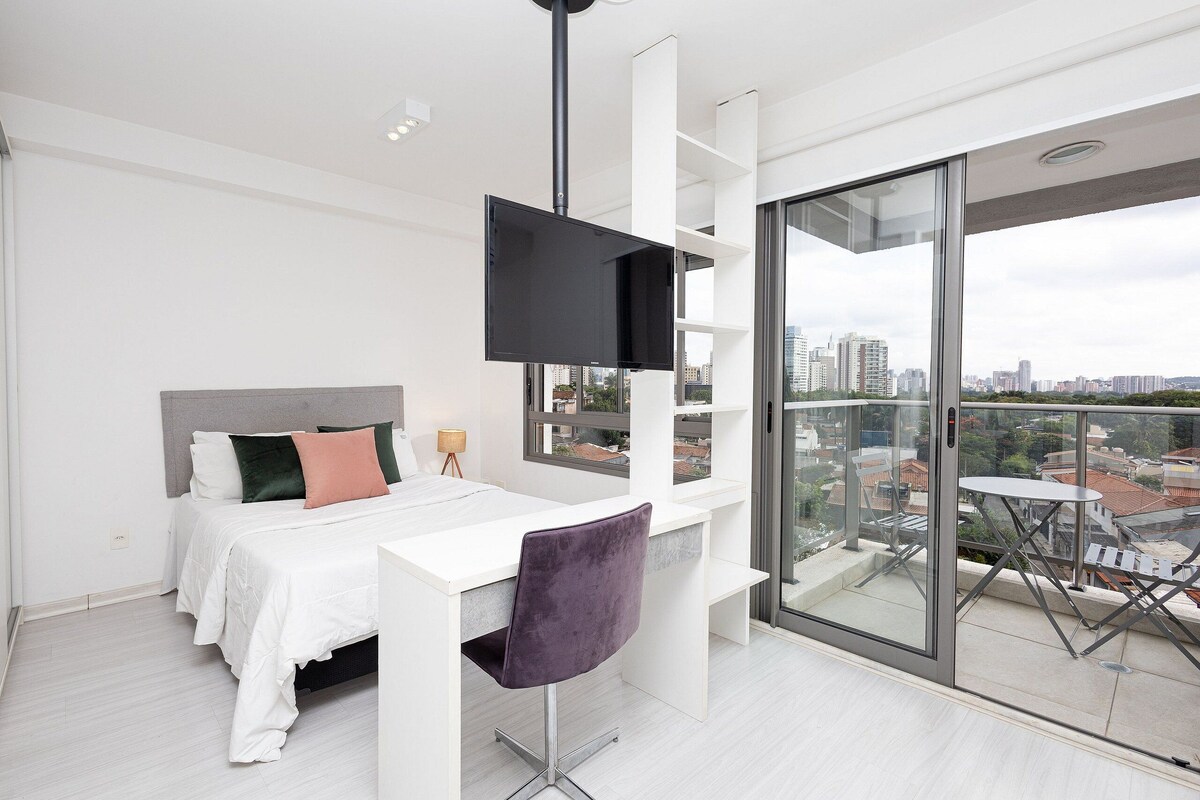 Quarto moderno com cama, escrivaninha, cadeira, TV e varanda com vista para a cidade.