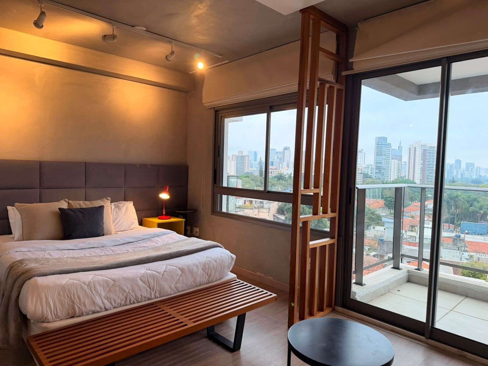 A modern bedroom with a large bed, a bedside table with a lamp, a bench, and a view of a cityscape through large windows.