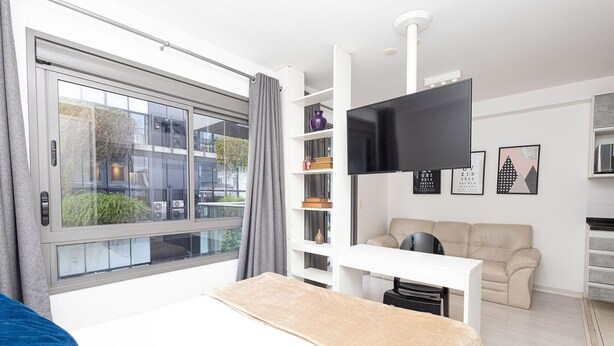 A modern bedroom with a large bed, a TV mounted on the wall, a sofa, and a window with curtains. - Vossa Bossa Vila Madalena Diseño (São Paulo)