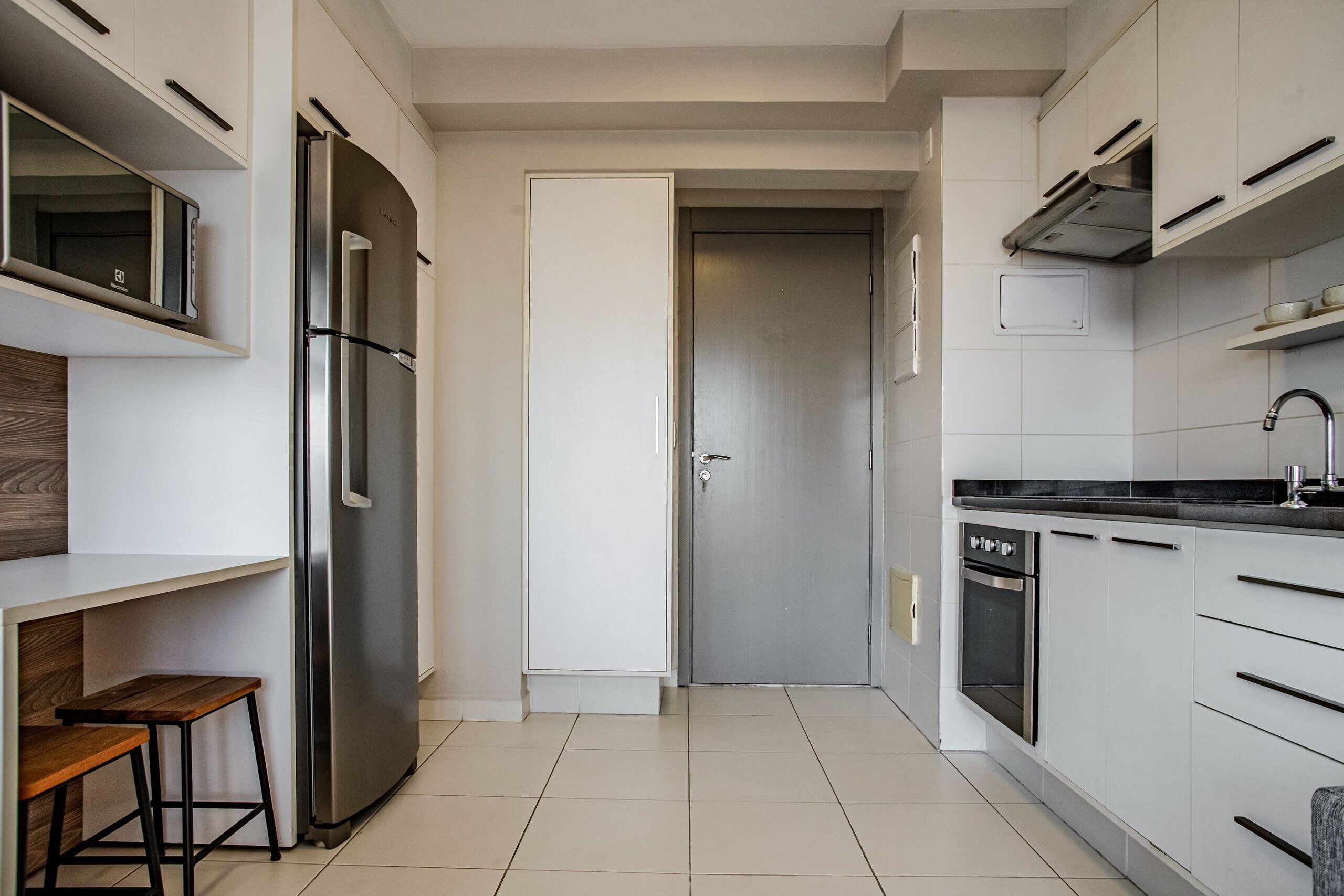 A modern kitchen with white cabinets, a stainless steel refrigerator, a built-in oven, and a wooden stool.