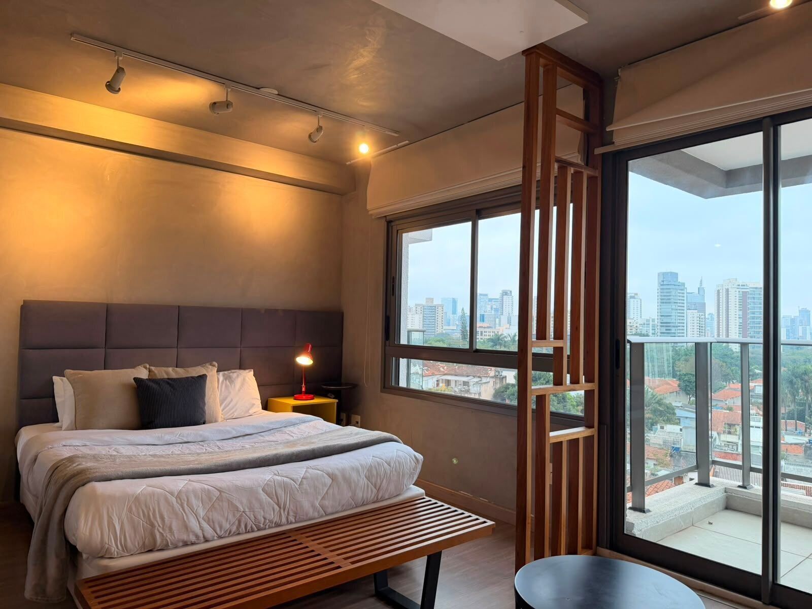 Una camera da letto moderna con un letto grande, un comodino con una lampada, una panca e vista sulla città attraverso ampie finestre.