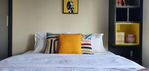 A neatly made bed with white bedding and colorful pillows, a framed artwork on the wall, and a bookshelf with books and decorative items. - Vossa Bossa Vila Madalena Diseño (São Paulo)