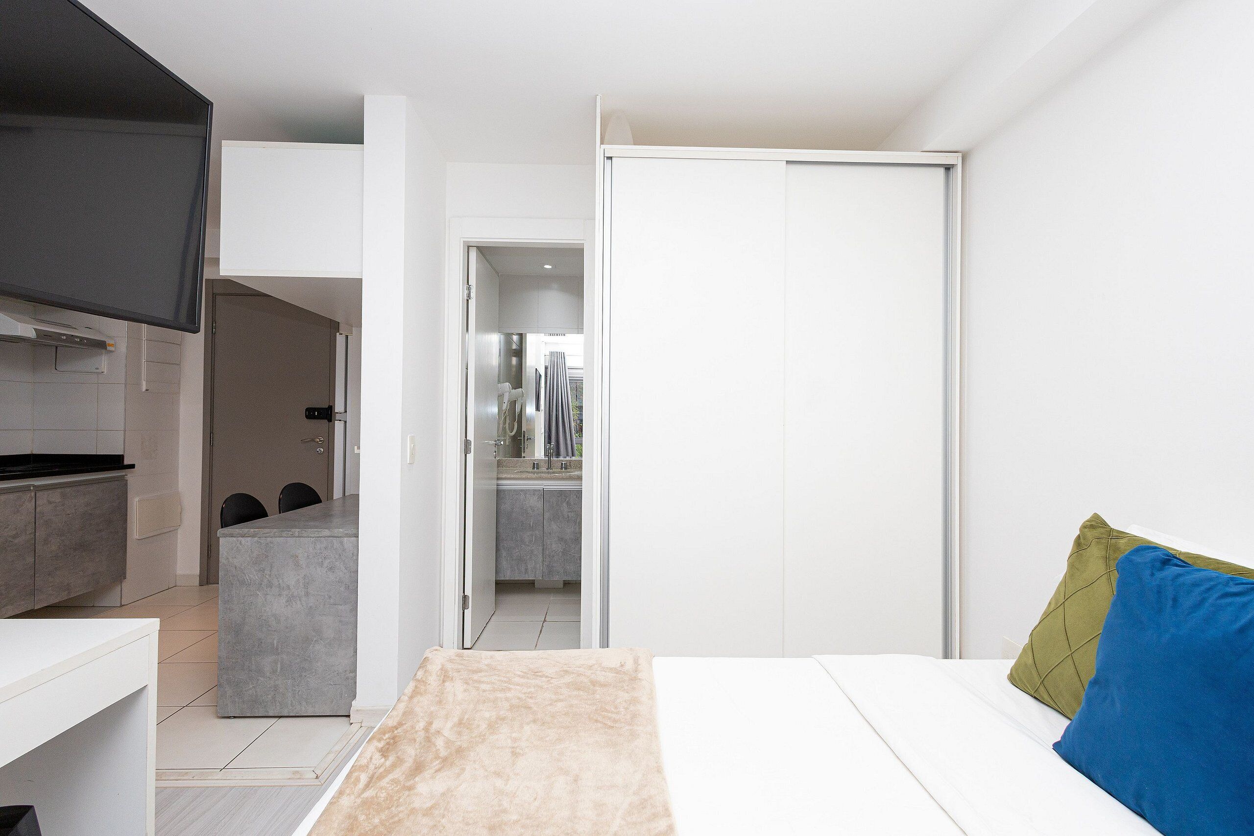 A modern bedroom with a bed, a TV, and a kitchenette in the background.