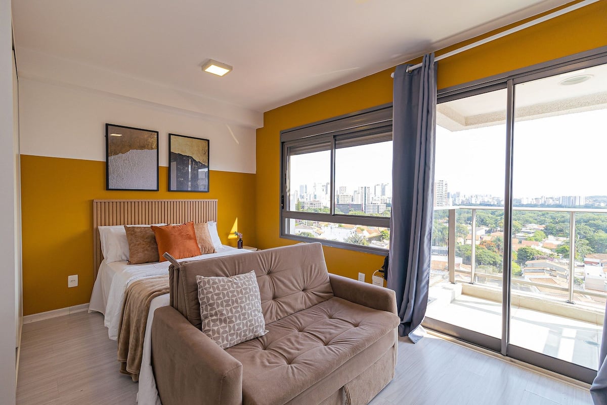A modern bedroom with a large window, a bed, a sofa, and two framed artworks on the wall.