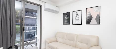 A modern living room with a beige sofa, a glass door leading to a balcony, and three framed pictures on the wall.