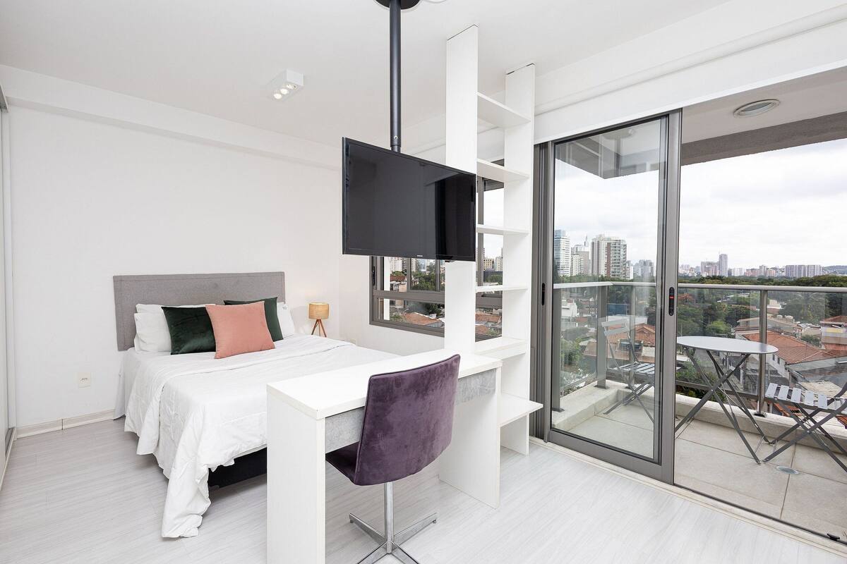 A modern bedroom with a bed, a desk, a chair, a TV, and a balcony with a city view.