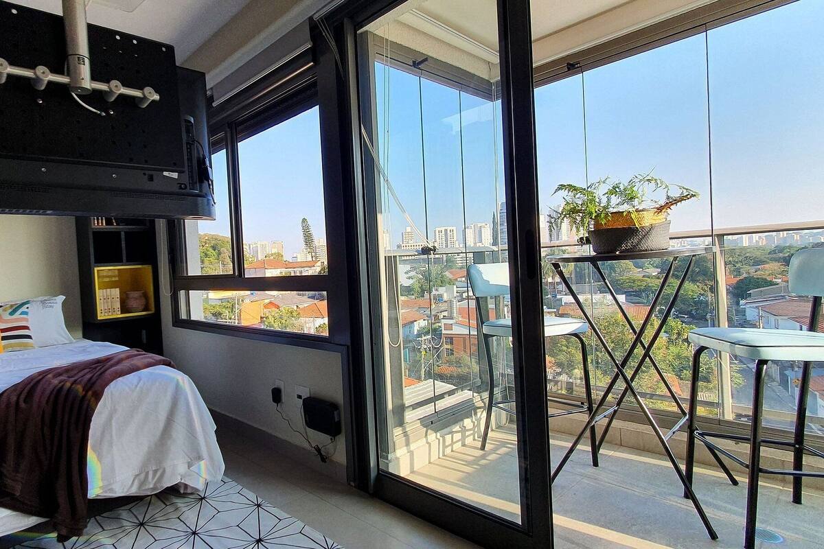 A modern bedroom with a bed, a TV mounted on the wall, and a balcony with a chair and a potted plant.