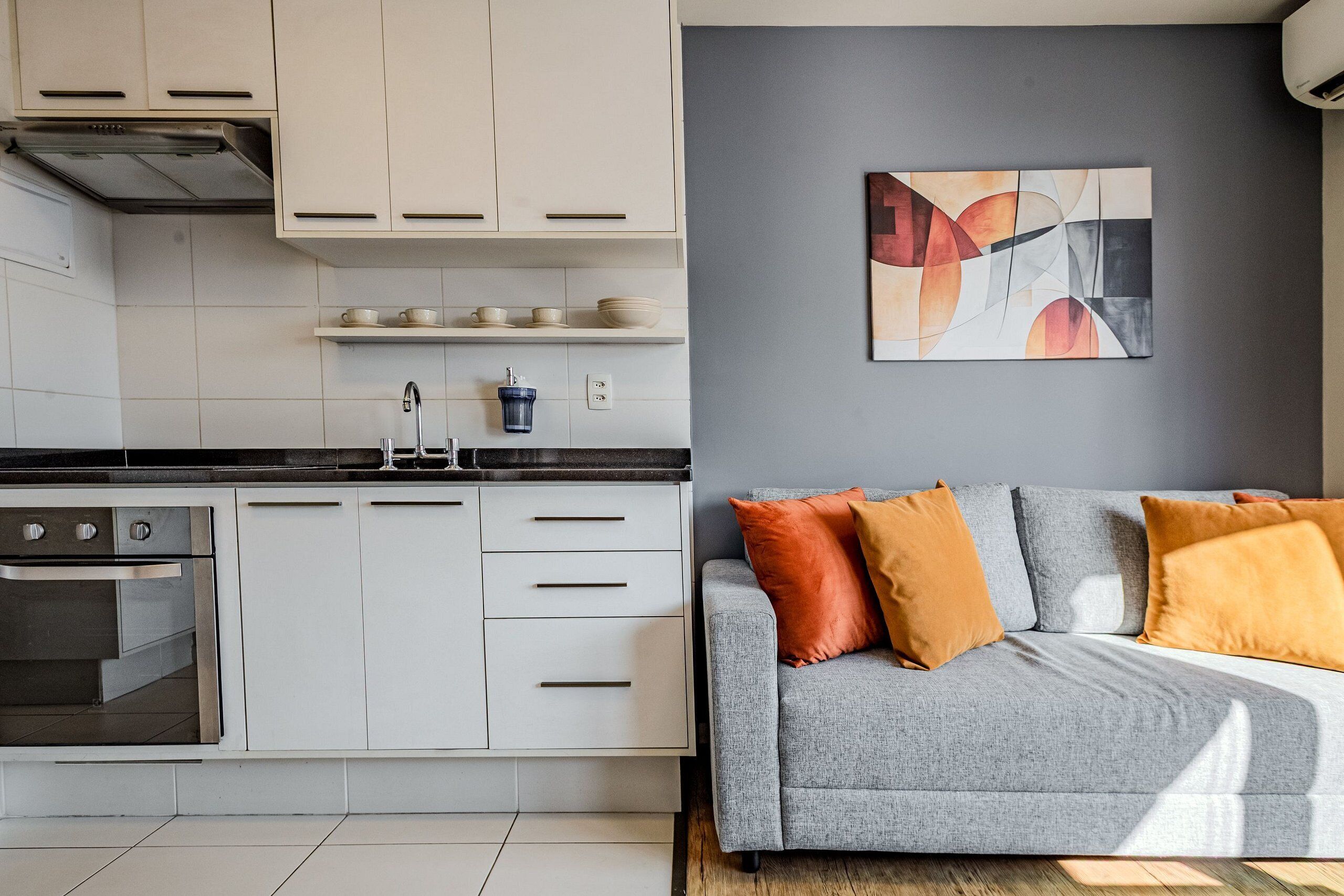 Una cocina moderna con armarios blancos, horno integrado y encimera negra. En la sala de estar, hay un sofá gris con cojines anaranjados y una obra de arte abstracta en la pared.