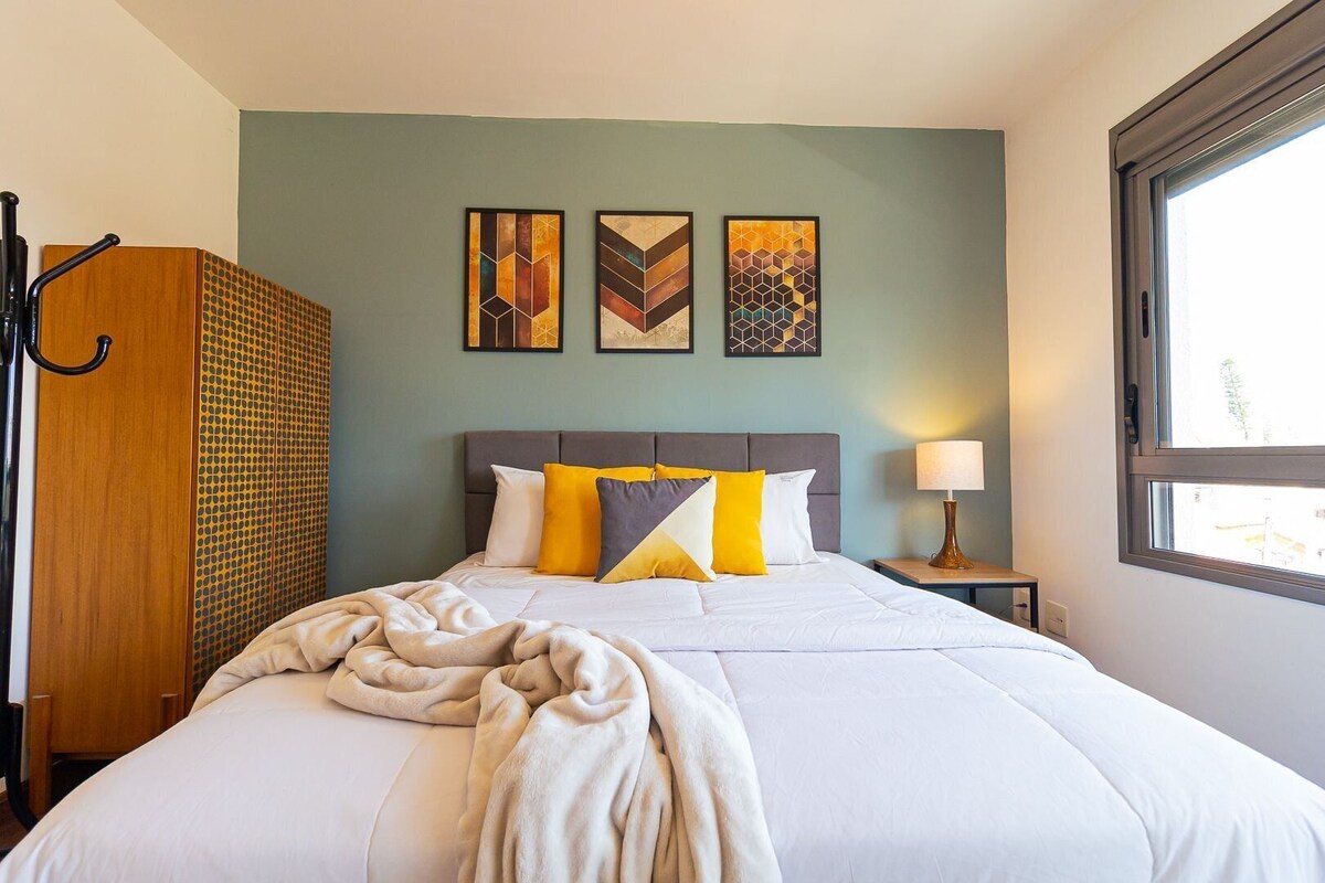 Quarto de hotel com cama, mesa de cabeceira, abajur e obras de arte na parede.