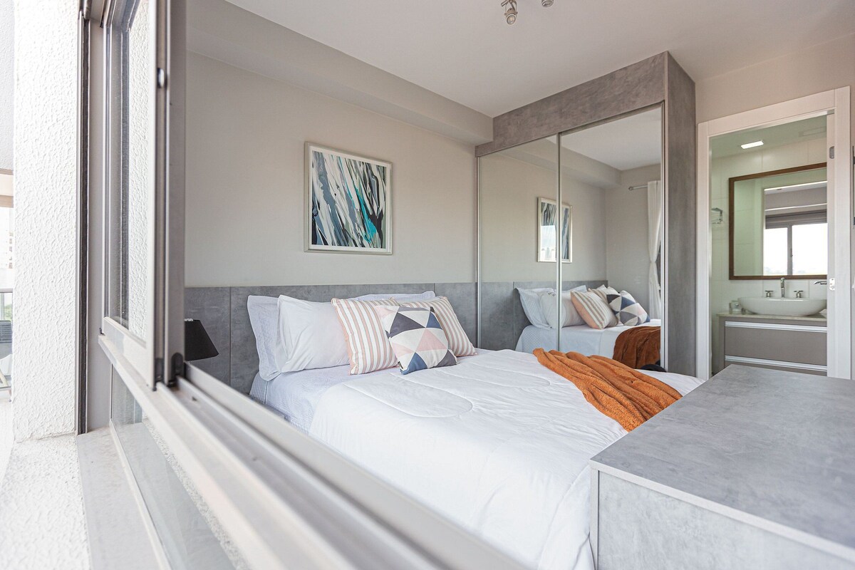 A modern bedroom with a large bed, a bedside table, a mirror, and a bathroom visible through an open door.