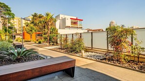A modern outdoor patio area with a wooden bench, a small garden with various plants, and a white building with a red accent. - Vossa Bossa Vila Madalena Diseño (São Paulo)
