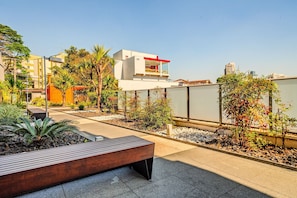 A modern outdoor patio area with a wooden bench, a small garden with various plants, and a white building with a red accent. - Vossa Bossa Vila Madalena Diseño (São Paulo)