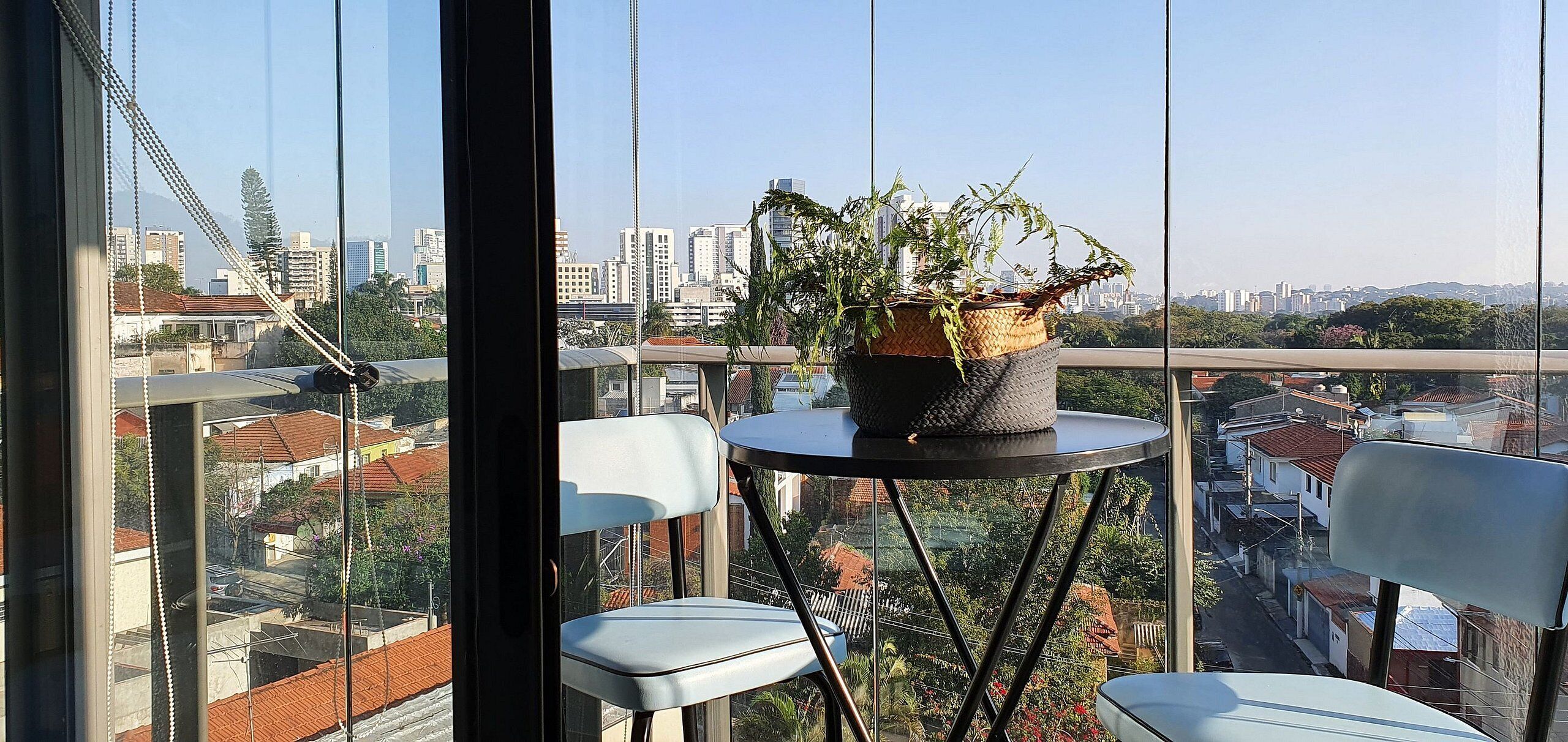 A balcony with a potted plant, two chairs, and a view of a cityscape including buildings and a bridge.