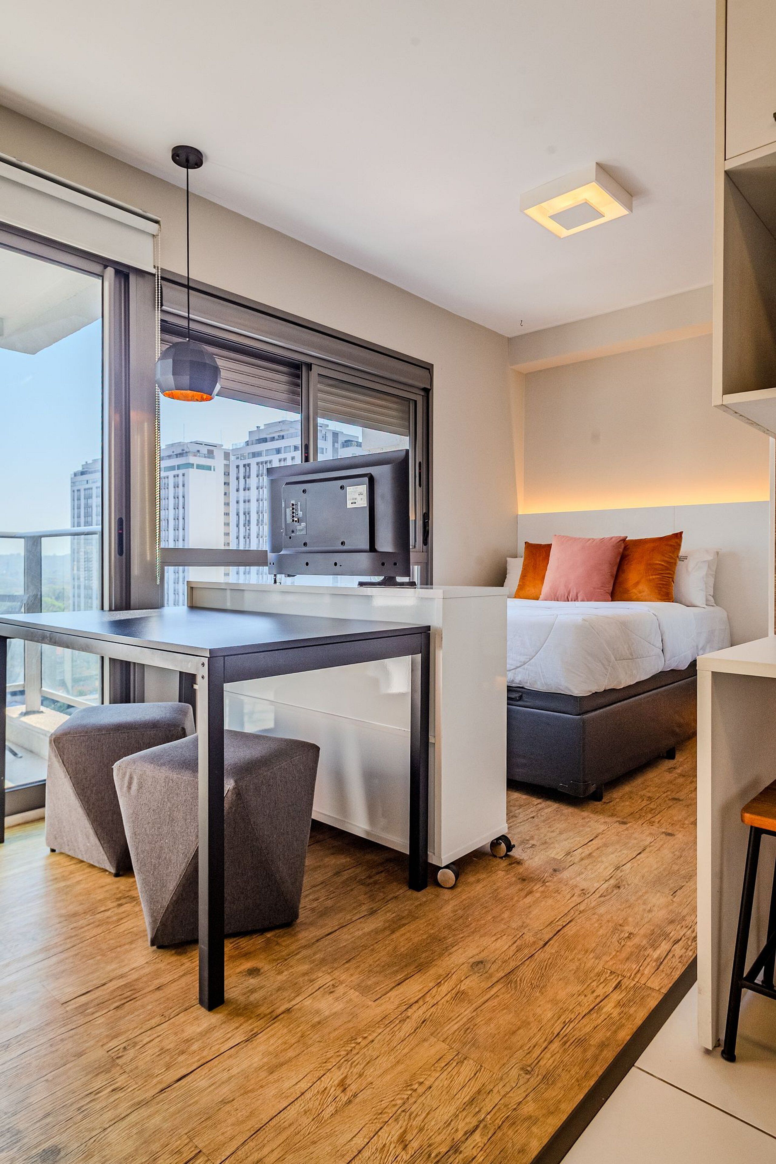 Een moderne hotelkamer met een groot raam, een bureau met een computer, een bed met kussens en een klein tafeltje.