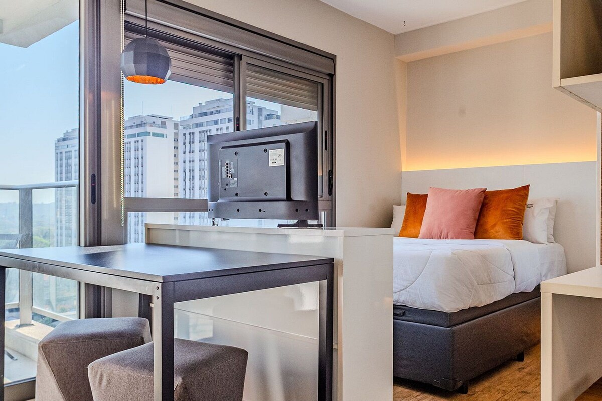 A modern hotel room with a large window, a desk with a computer, a bed with pillows, and a small table.