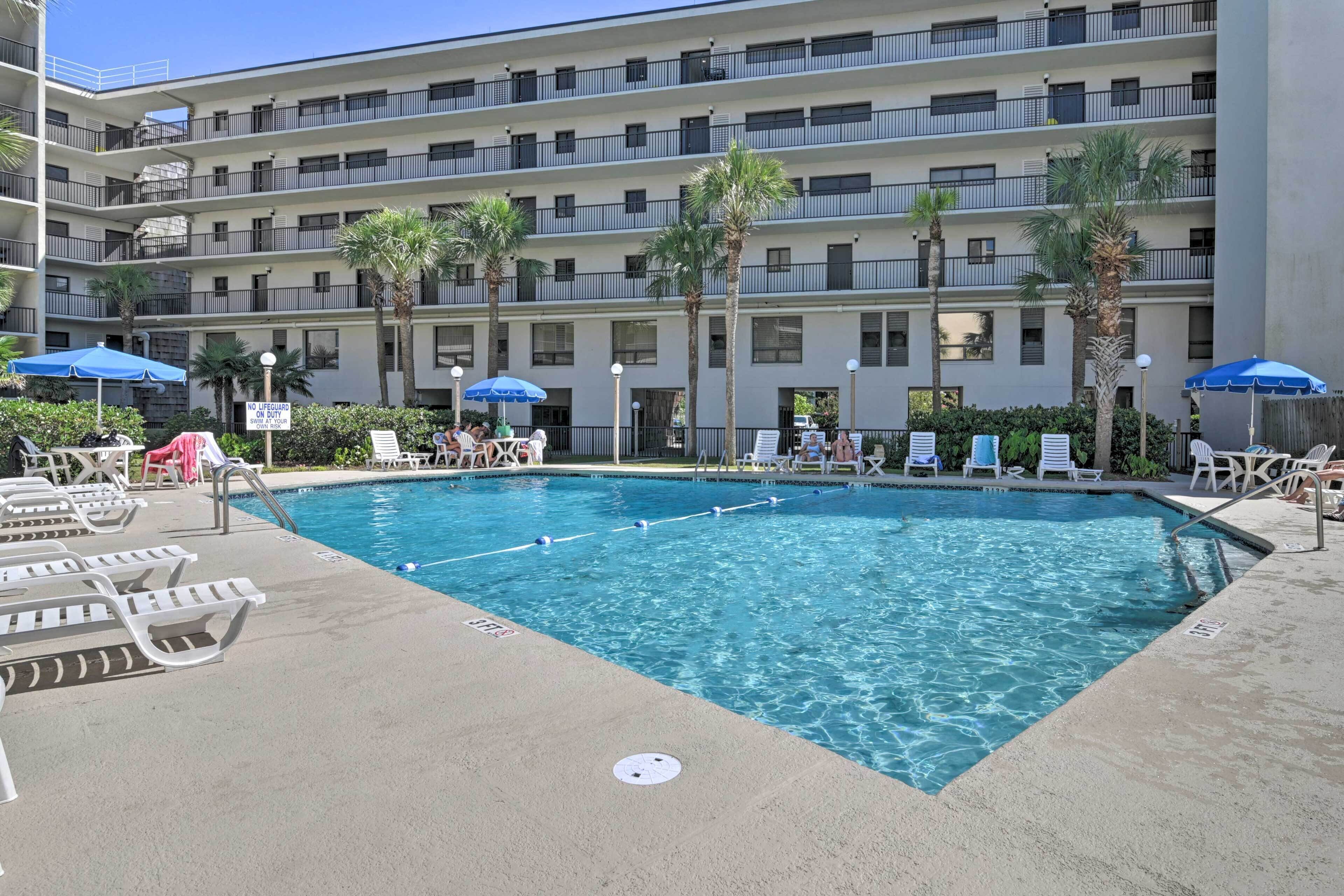 A hotel pool area with multiple lounge chairs, umbrellas, and a multi-story building in the background.
