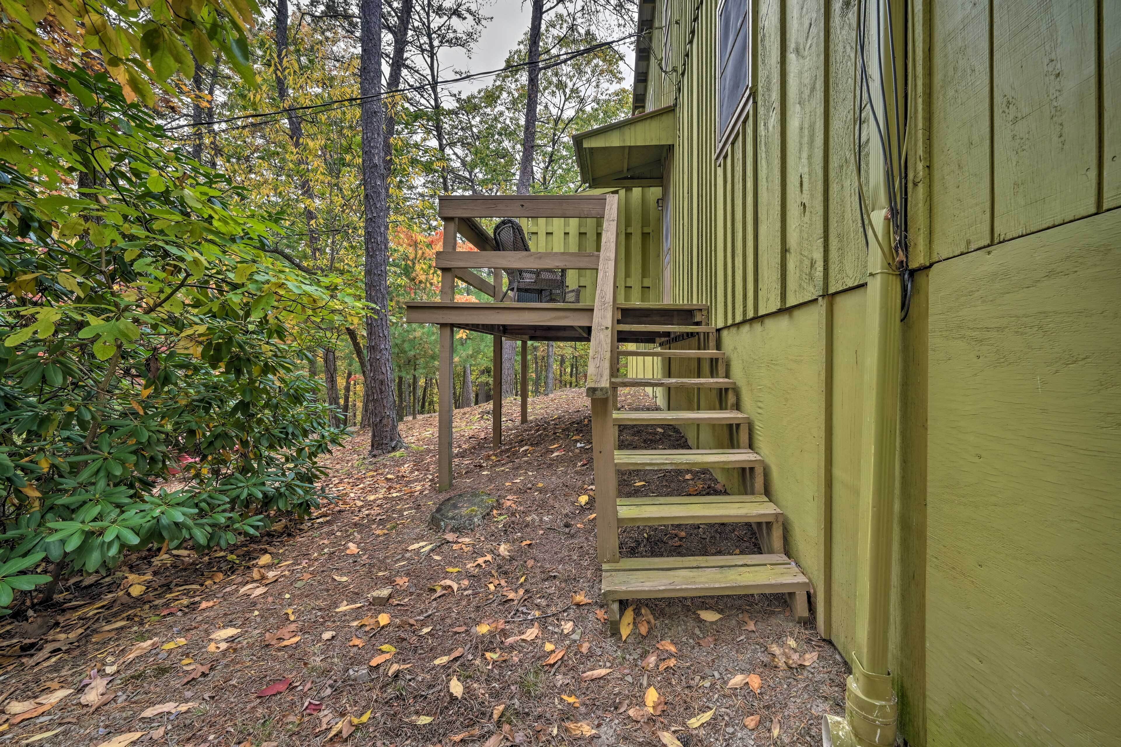 A wooden staircase leading to a deck with a chair, surrounded by trees and a green building.