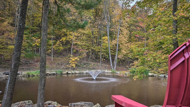 Ein ruhiger Teich mit einer Fontäne, umgeben von Bäumen mit Herbstlaub, und ein roter Stuhl im Vordergrund.