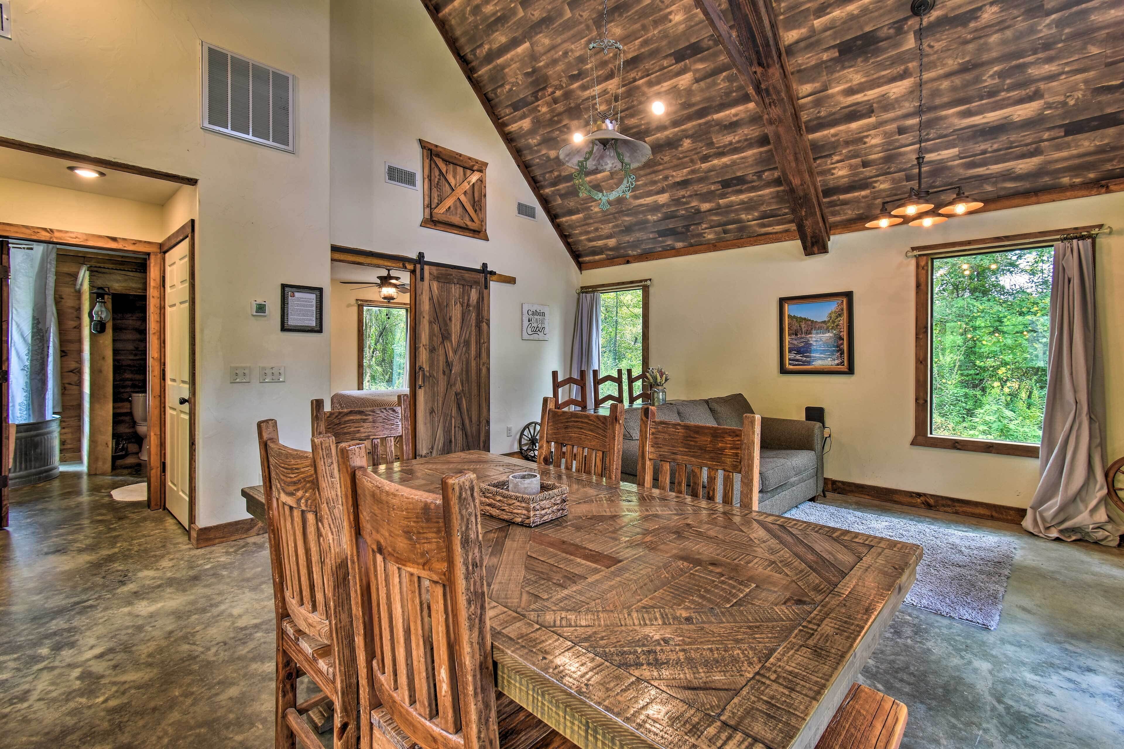 A spacious dining area with a large wooden table and chairs, a rustic ceiling, and large windows offering a view of the outdoors.