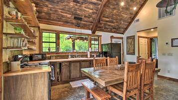 A rustic kitchen with wooden beams, a large dining table, and a view of greenery outside.