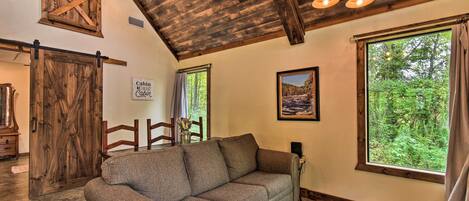 Interior acogedor de una cabaña con techo de madera, un sofá, una mesa de comedor y grandes ventanales que dan al bosque.