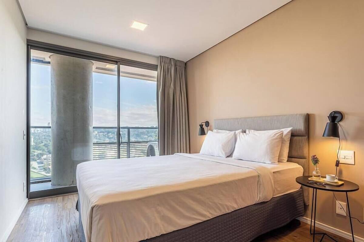 A modern bedroom with a large bed, a small bedside table, and a view of the city through a large window.