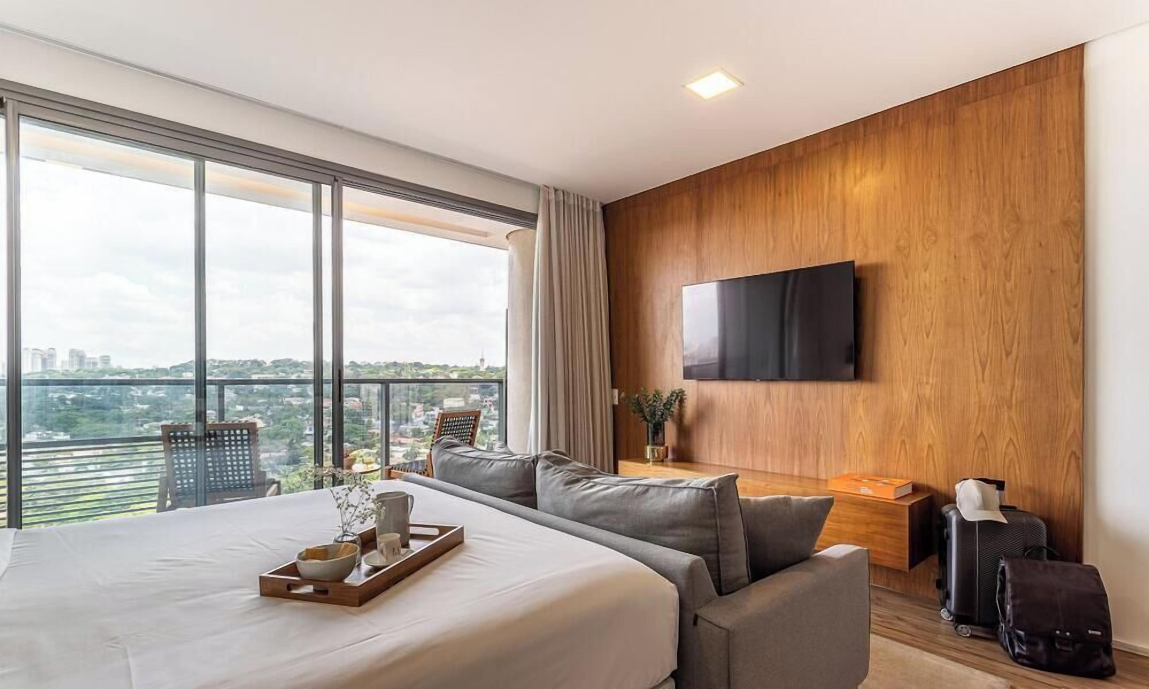 A modern hotel room with a large bed, a seating area, a TV mounted on the wall, and a view of the city.