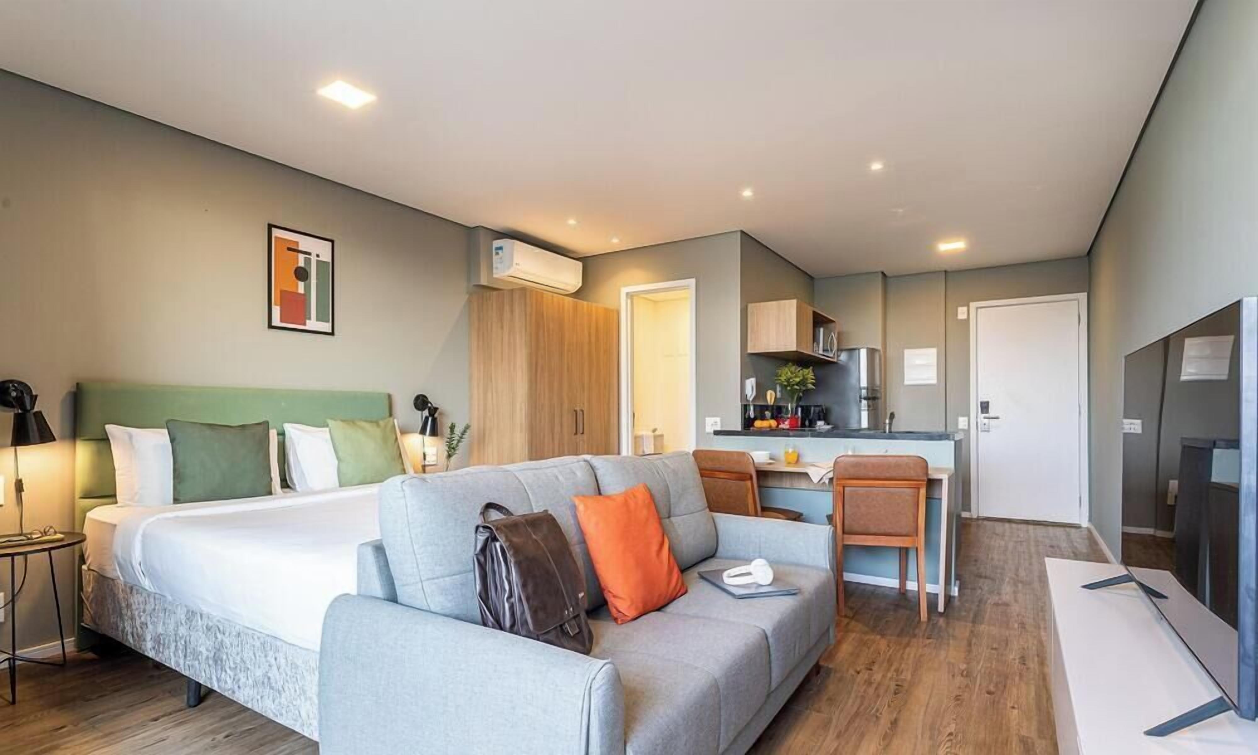 A modern hotel room with a bed, a sofa, a desk, and a kitchenette.