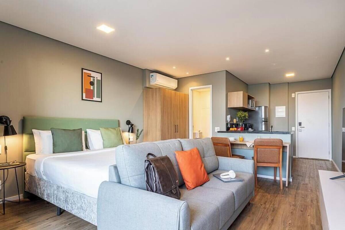 A modern hotel room with a bed, a sofa, a desk, and a kitchenette.